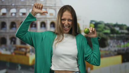 Young woman holding green lime with thumb to chest and raised arm in building coliseum wearing...
