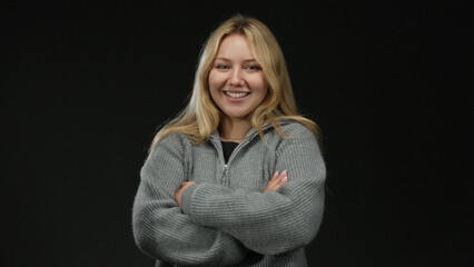 Young woman smiling confidently with arms crossed in front of a black background, wearing a cozy...