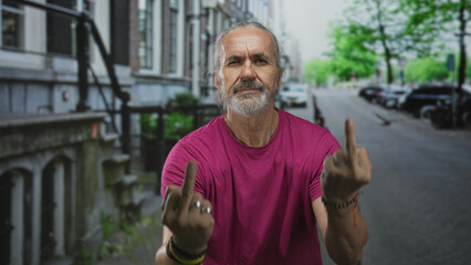 Man shows both middle fingers on a city street, wearing magenta shirt and long grey hair tied back...