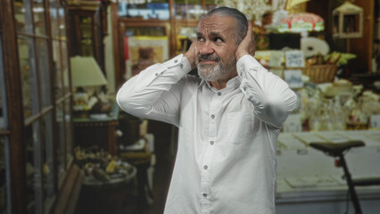 Man with hoary long hair and hands to ears amid a cluttered antique shop building filled with lamps...