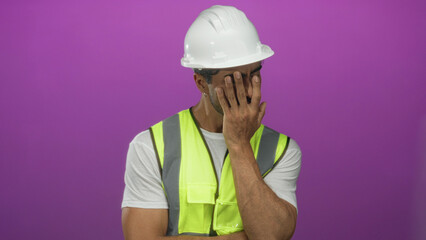 Hispanic man wearing white hardhat and reflective vest covers face with hands in purple studio;...