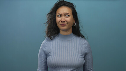 Obraz premium Woman points finger to temple in blue studio with puzzled expression and raised eyebrow; confusion thinking doubt.