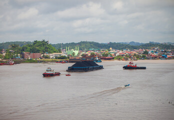 Obraz premium Contrasting activity on the Mahakam River in Samarinda, East Borneo, Indonesia. A massive coal barge, guided by tugboats, passes a small local motorboat, showcasing the river's diverse traffic