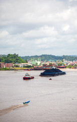 Contrasting activity on the Mahakam River in Samarinda, East Borneo, Indonesia. A massive coal barge, guided by tugboats, passes a small local motorboat, showcasing the river's diverse traffic