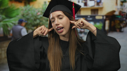 Woman in black graduation gown and cap plugs ears with fingers on a vibrant city street lined with...
