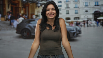 Young hispanic woman wearing tank top with hands on chest on city street with blurred building behind her; gratitude.