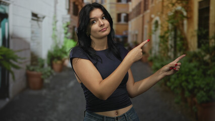 Woman with exposed navel points finger to side on sunlit street between pastel buildings; curiosity.