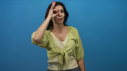 Young hispanic woman standing smiling makes ok sign around her eye in bright blue studio setting; approval.