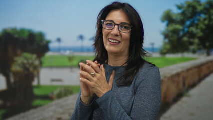 Fototapeta premium Woman wearing glasses smiling with clasped hands amid green grass in park pathway by stone wall; serenity.