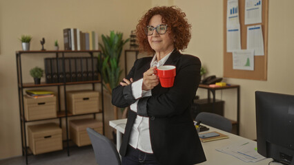 Woman standing in office holding red cup wearing glasses and blazer surrounded by shelves and plants in a modern workplace setting.