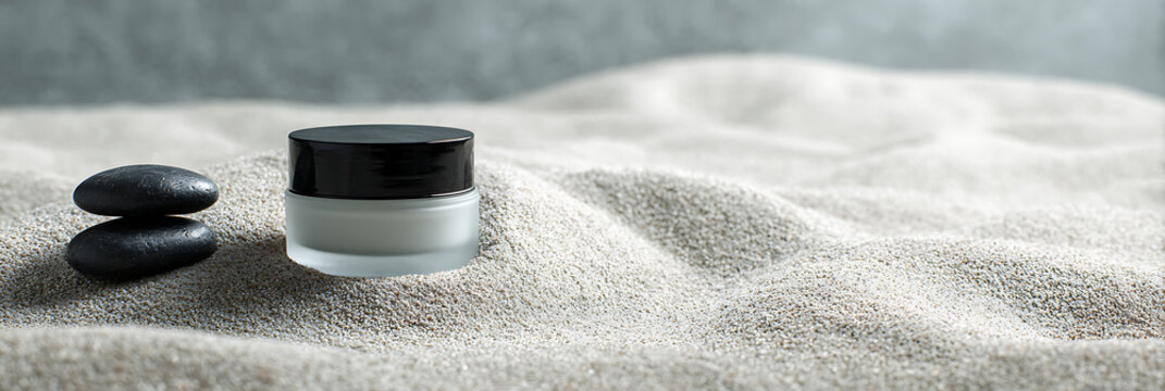 Minimalist composition featuring a frosted glass cosmetic cream jar and balanced black zen stones resting on soft, rippled white sand, emphasizing natural skincare.