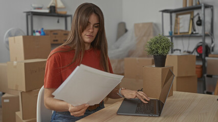 Woman examining documents in living room surrounded by packing boxes and laptop signifies moving,...