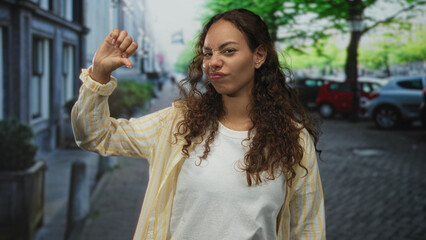 Fototapeta premium Young african american woman showing thumbs down with hand raised on city street near parked cars and trees, casual shirt and neutral expression; disapproval rejection critique.