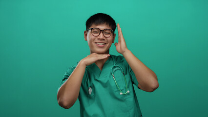 Young chinese man in green scrubs with stethoscope smiling against isolated green background wall...