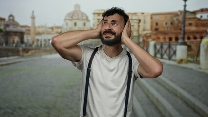 Fototapeta premium Hispanic man with beard looking distressed in old town rome holding his head in hands wearing suspenders against soft focus background of historical architecture