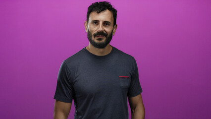 Naklejka premium Man standing against a vibrant pink background, wearing a casual grey shirt, exuding confidence and calmness, his dark beard and hair complementing the bright setting.