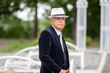 An elderly man in a stylish suit and hat poses in the garden on a sunny day