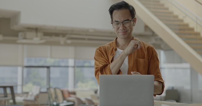 Senior Asian businessman wearing headset celebrates success during online meeting in modern co-working space. Remote work and business communication concept