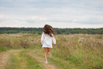 Girl runs along a grassy path in a field during a cloudy day surrounded by greenery and wildflowers