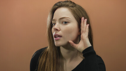 Fototapeta premium Woman with brown hair and serious expression cupping hand to ear against orange wall in studio; focused listening.