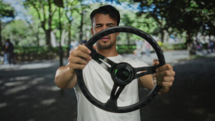 Obraz premium Young hispanic man in a park holding a steering wheel, expressing surprise against a green outdoor background.