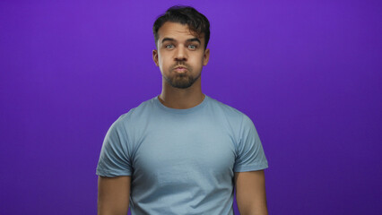 Obraz premium Young man inflates his cheeks and touches them with fingers in front of isolated purple background displaying playful expressive gesture.