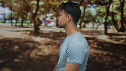 Young man in a blue shirt stands thoughtfully in a sunlit park surrounded by green trees, evoking a...