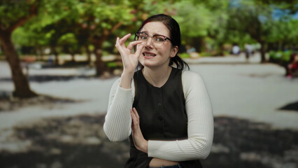 Fototapeta premium Woman with glasses gesturing outdoors in a sunny park, surrounded by vibrant green trees, expressing various emotions with an inquisitive and thoughtful demeanor.
