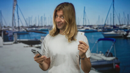 Fototapeta premium Man holding smartphone and creditcard while looking at phone beside docked boats on street pier; convenience.
