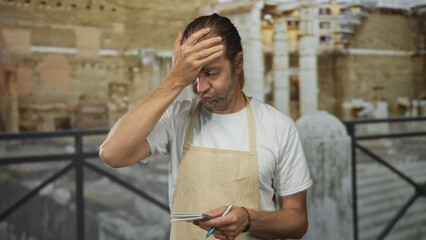 Man with blond hair and apron holds notebook and touches forehead in building among ancient ruins;...