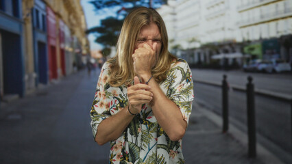 Fototapeta premium Blond man with long hair covers nose with hand on city street sidewalk near bollards amid empty storefronts; disgust.