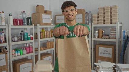 Young hispanic man holding paper bag in building surrounded by donation boxes and metal shelves,...