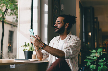 Bearded man holding smartphone in hand near cafe window, casual freelancer with coffee, concept of modern digital interaction and everyday communication.