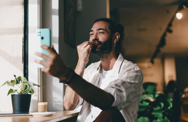 Smiling man sending air kiss into smartphone during video call, casual freelancer with coffee in cafe, digital lifestyle concept of communication and modern connection.