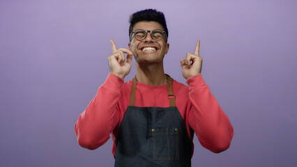 Young hispanic man in glasses and apron points upward smiling against a solid purple background, wearing a red sweater, expressing joy and positivity in a studio portrait.