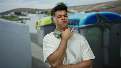 Fototapeta premium Young man with headphones poses on a city street expressing frustration near recycling bins surrounded by urban scenery and white architecture enhancing the outdoor atmosphere.