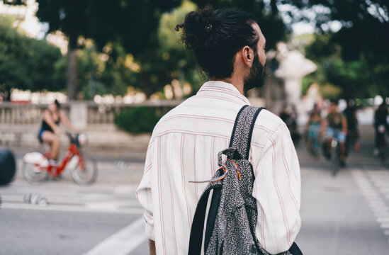 Man with backpack stands at crosswalk, viewed from behind, looking at street activity with calm curiosity, symbolizing mindful movement, solo exploration and modern urban lifestyle.