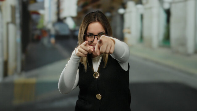 Young blonde woman in glasses pointing playfully in an outdoor street setting, embodying a cheerful and engaging atmosphere.