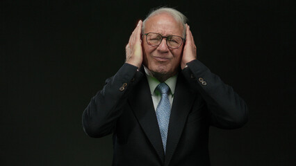 Senior man in business suit covers ears against isolated black background, expressing discomfort or frustration with eyes closed, illustrating a powerful human emotion.