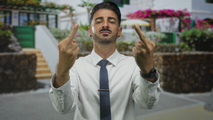 Young hispanic man in white shirt and tie flips both middle fingers toward camera on sunny city...