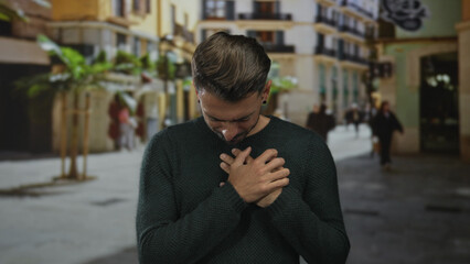 Young hispanic man wearing dark green sweater holds hands on chest with bowed head on narrow...