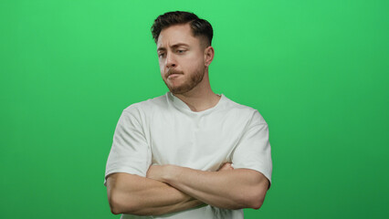 Young man with beard and white shirt stands thoughtfully against isolated green background wall posing with folded arms in contemplative attitude.