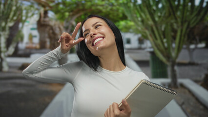 Woman smiling and posing with a notebook in an outdoor park setting with trees, showcasing joy and positivity on a sunny day.