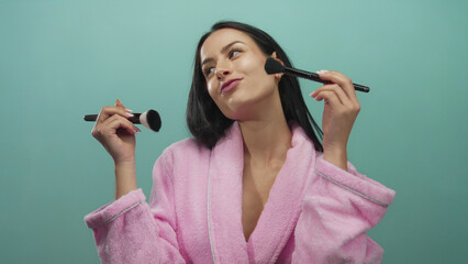 Obraz premium Woman applying makeup with brushes on an isolated green background wearing a pink robe, exuding beauty, femininity, and self-care in a vibrant yet calm setting.