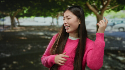 Fototapeta premium Woman in a pink sweater gestures playfully outdoors in a park with trees, smiling and enjoying a sunny day.