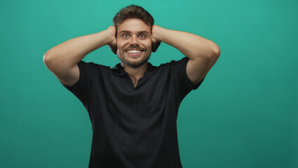 Fototapeta premium Young hispanic man with hands over ears posed in black polo in teal studio under studio lighting; surprise reaction.