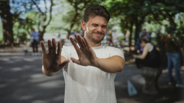 Young hispanic man with palms out and hands raised in park to stop close contact; disgust avoidance.