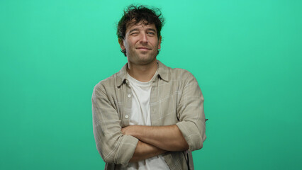 Fototapeta premium Man with arms crossed standing confidently against a vibrant green background, exuding charm and warmth in a casual style.