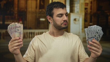 Young hispanic man smiles and holds saudi riyal and dollar banknotes in church building; prosperity...