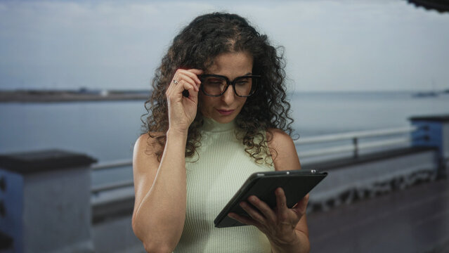 Woman touching tablet with exposed shoulder and curly hair on seaside street; digital concentration. - Powered by Adobe